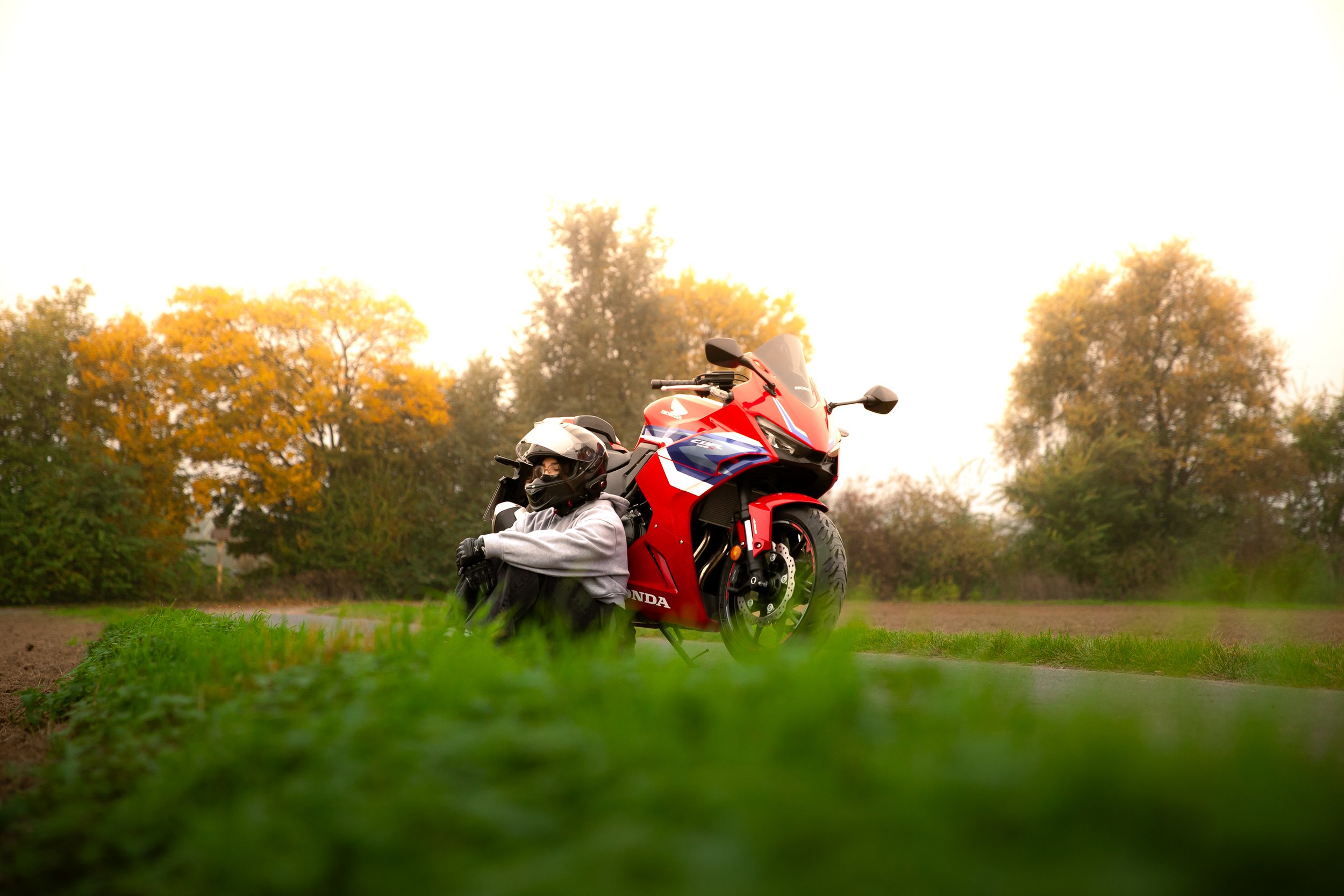 Mensch mit Helm sitzt am Boden neben rotem Motorrad in einem Feld, im Hintergrund Bäume mit herbstlichen Blättern.