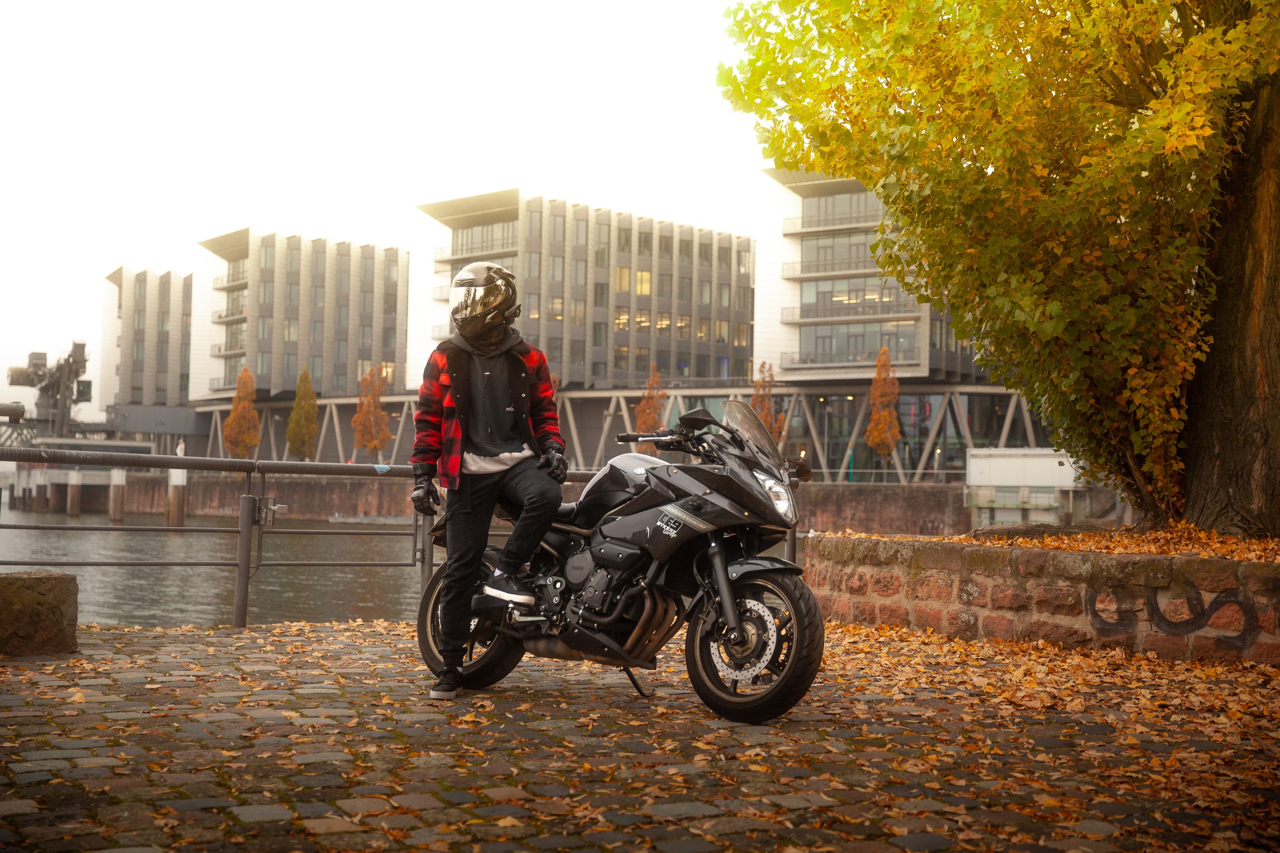 Motorradfahrer mit Helm und schwarzer Kleidung neben einer schwarzen Motorräder am Ufer bei herbstlichem Laub und modernen Gebäuden im Hintergrund.