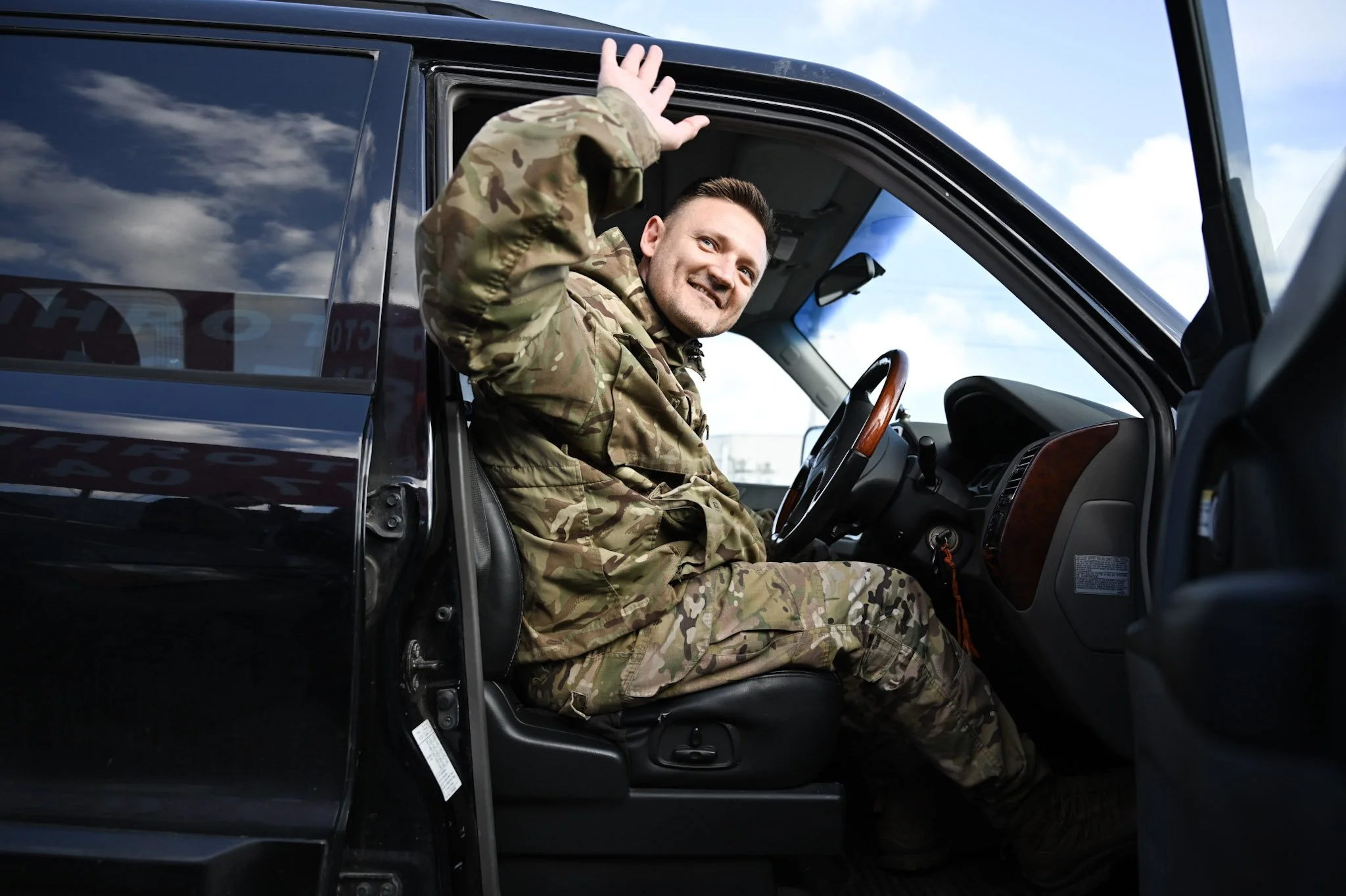 A man in camouflage military uniform sitting in the driver's seat of a black vehicle, smiling and waving through the open window at the sky with clouds.