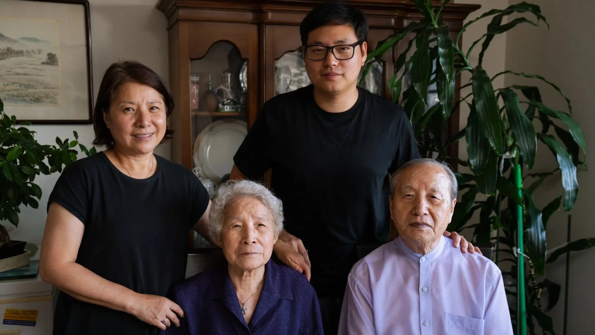 Family portrait with three elderly individuals seated and two middle-aged individuals standing behind them in a home setting with plants and artwork in the background.