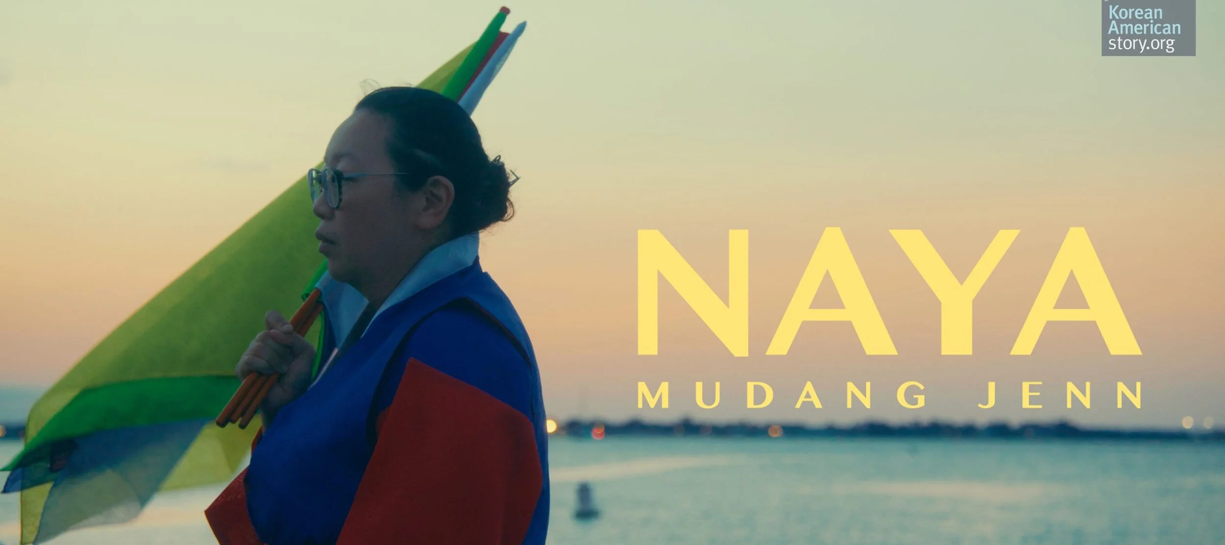 A person with glasses and dark hair pulled back is holding a multicolored flag and a bunch of sticks, standing by the water at sunset. The text reads "NAYA MUDANG JENN" and a small logo in the top right corner says "Korean American story.org."
