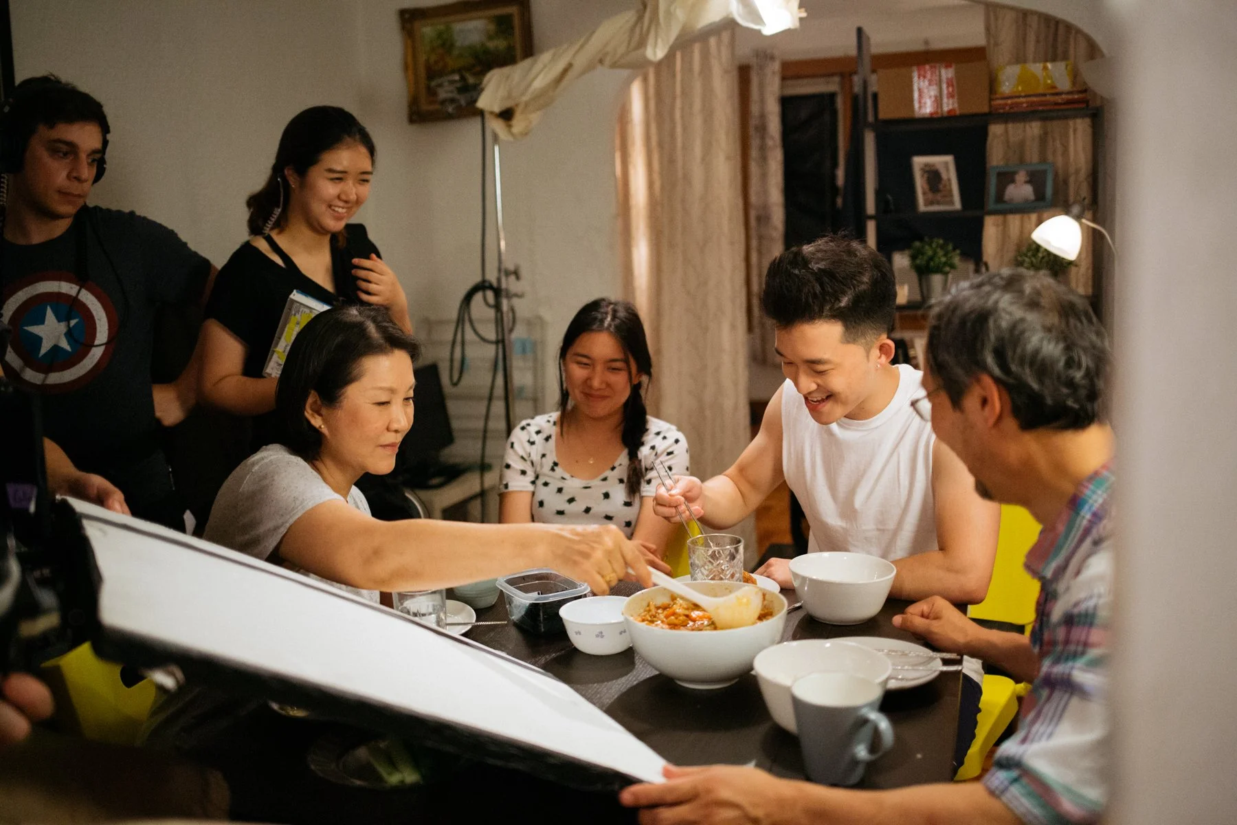 Family gathered around a table enjoying a meal together in a cozy home setting, with several bowls of food and drinks on the table.