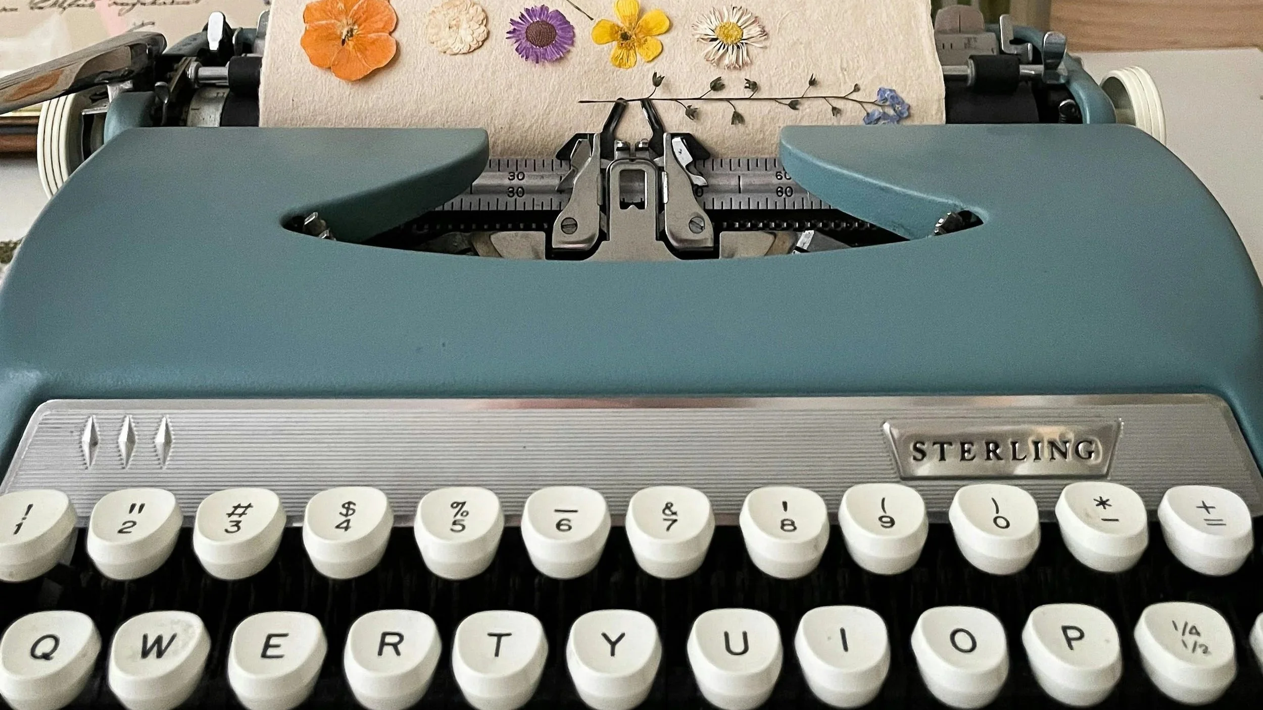 A vintage typewriter with a beige paper inserted into it, decorated with pressed flowers including orange, white, purple, yellow, and blue blossoms, with a ruler guide beneath the paper.