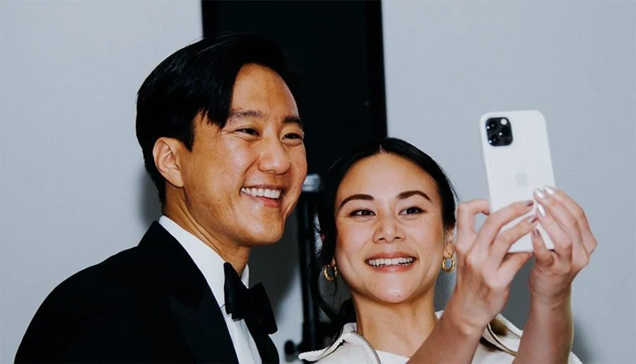 A man in a tuxedo and a woman in a white outfit taking a selfie together.
