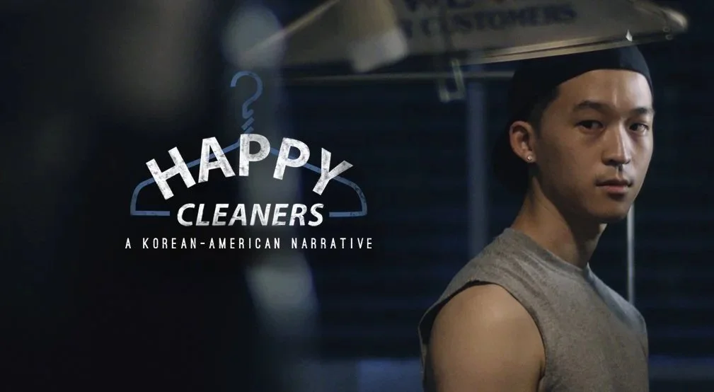A young man in sleeveless shirt and black cap standing in front of closed blue shutters at night, with text overlay reading "Happy Cleaners, A Korean-American Narrative."