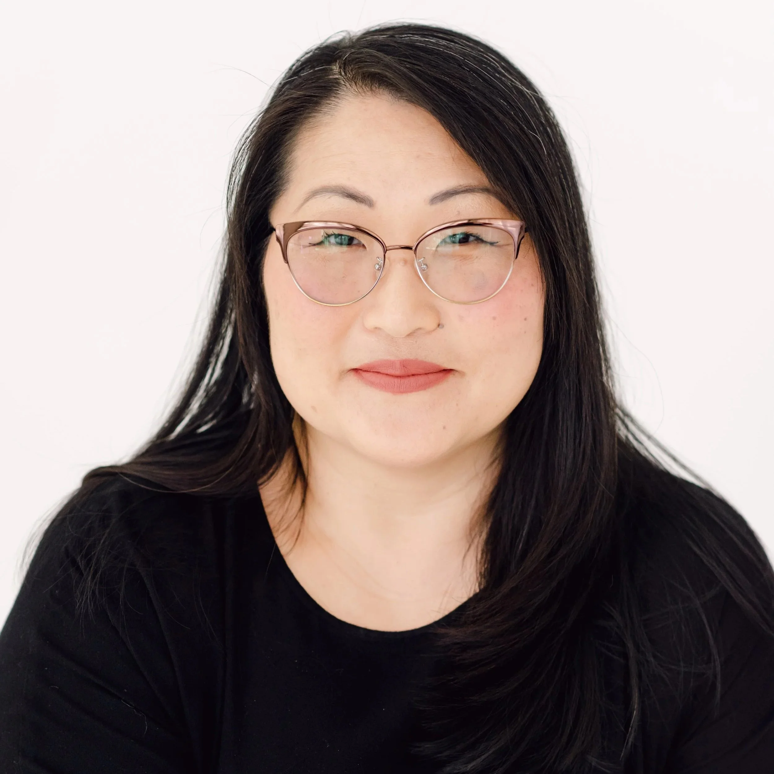 A close-up of a woman's face showing her forehead, eyes, and nose, with long black hair and wearing pink-rimmed glasses, against a plain white background.