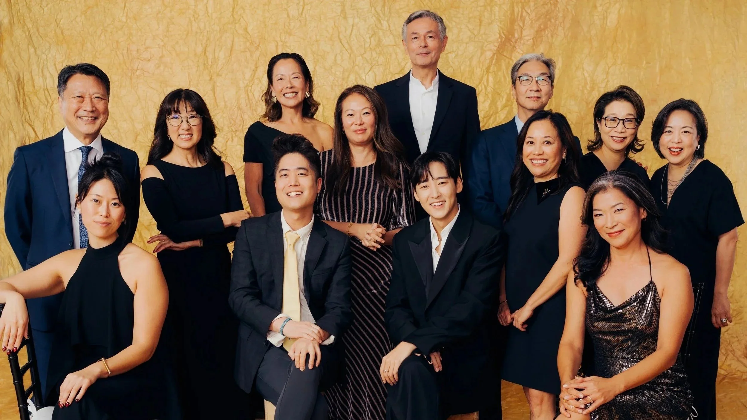 A diverse group of fifteen professionally dressed people posing together for a photo in front of a gold textured backdrop, smiling and standing close.
