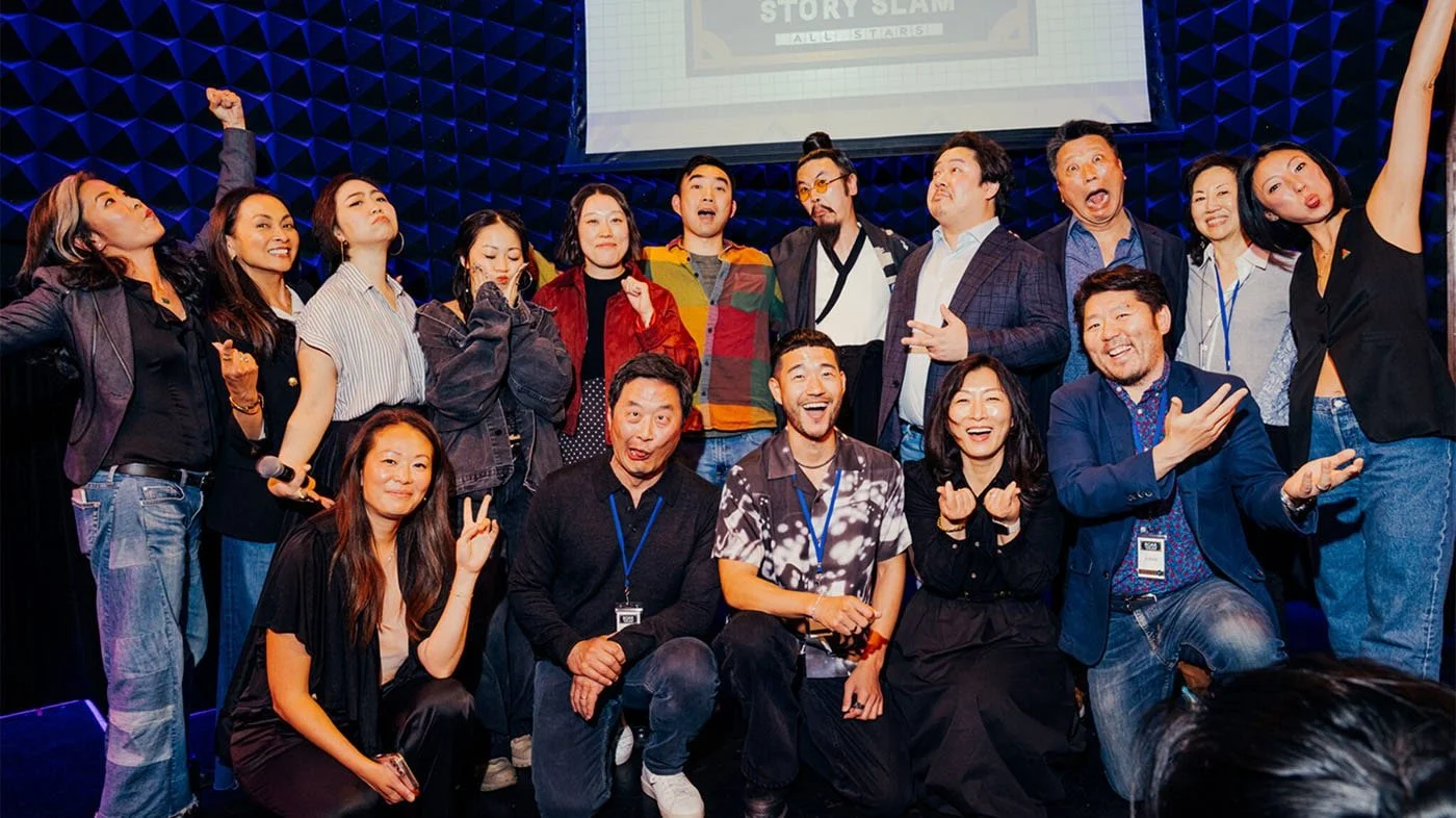 Group of smiling and expressive diverse people posing on stage during an event, some making gestures, background includes a large presentation screen and dark textured wall.