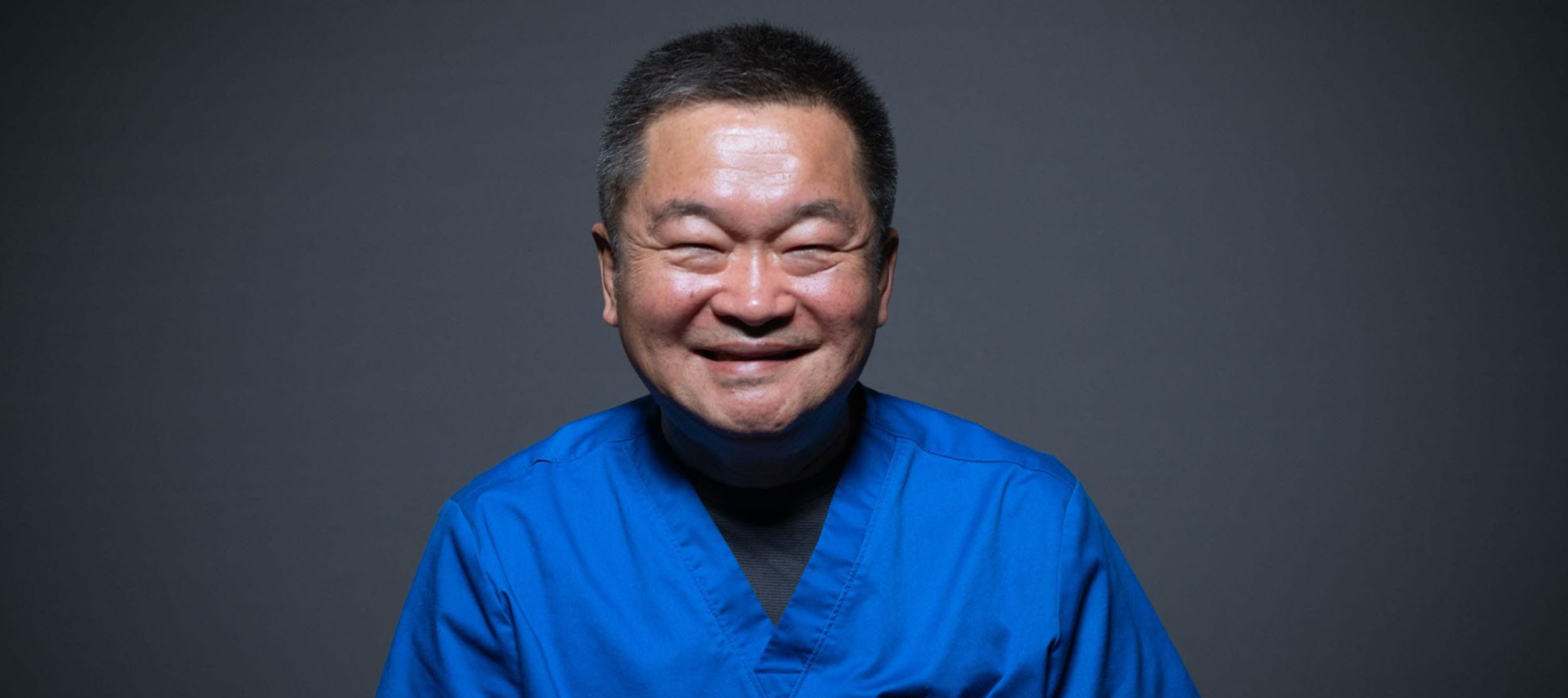 A person with short dark hair wearing a blue medical uniform and black turtleneck, smiling against a plain dark background.