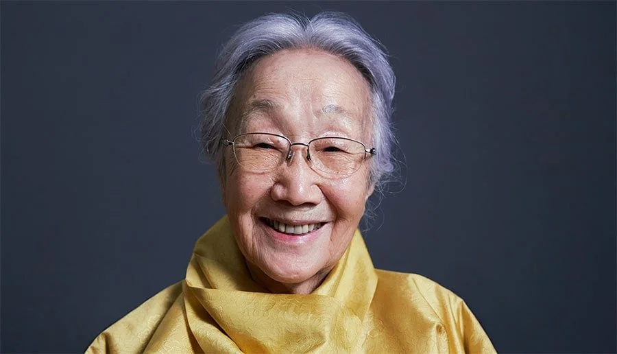 A smiling elderly woman with glasses and short gray hair, wearing a yellow garment, against a dark background.