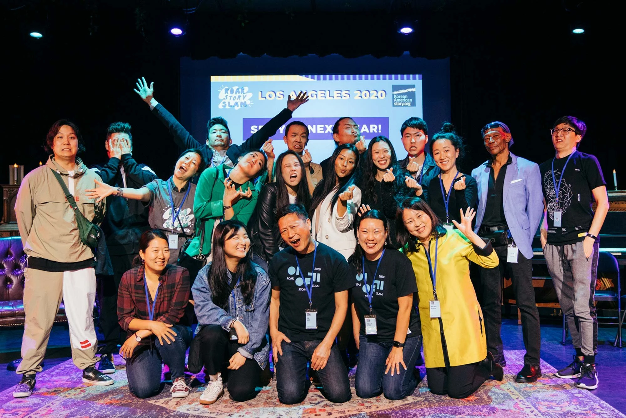 Group of people on stage at a Korean American storytelling event in Los Angeles 2020, posing for a photo with some making hand gestures, smiling, and standing in front of a screen with event details.
