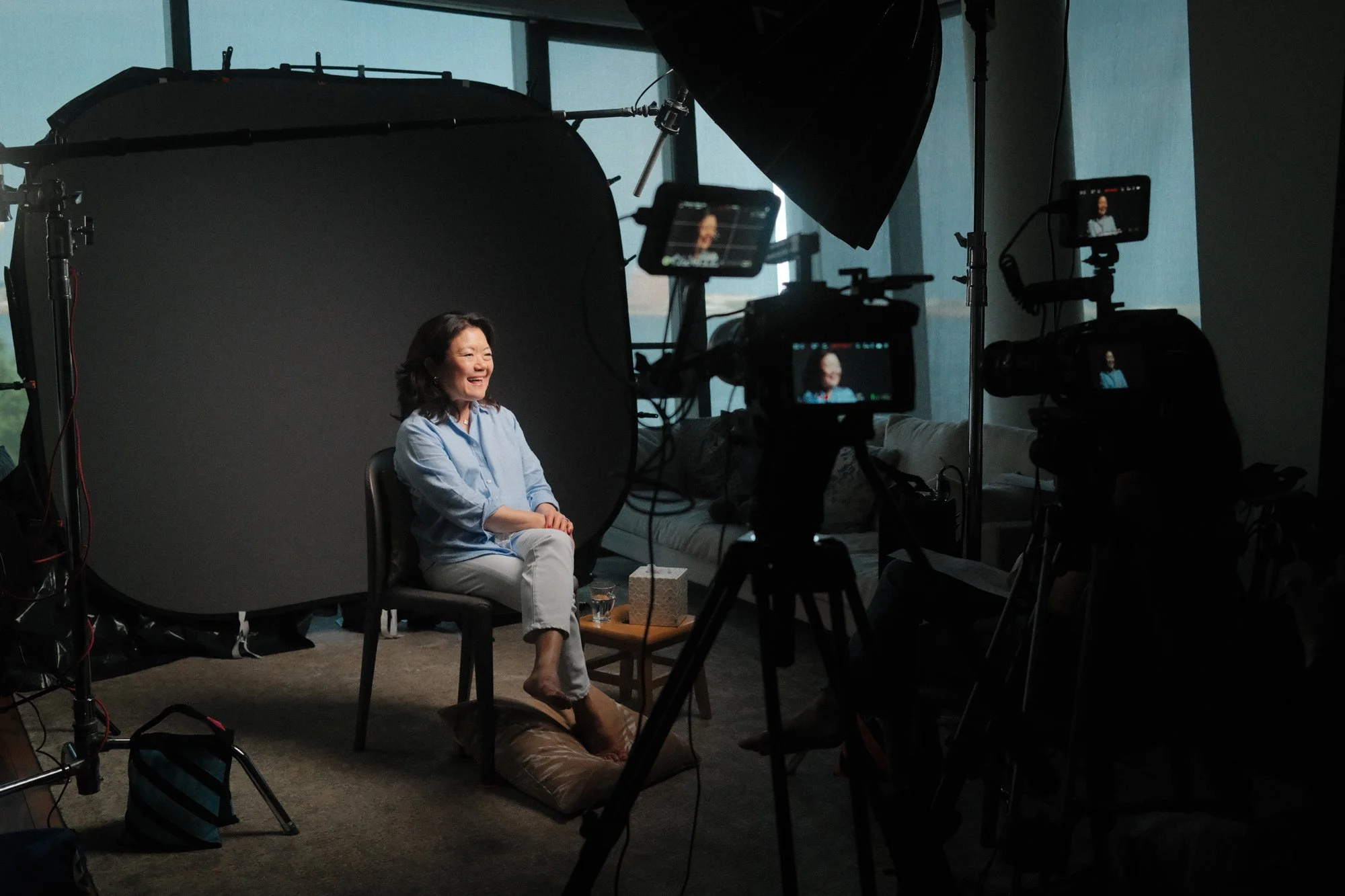 A woman with dark hair smiling and sitting on a chair in a photography studio, with professional lighting and camera equipment around her, and a large black reflector nearby.