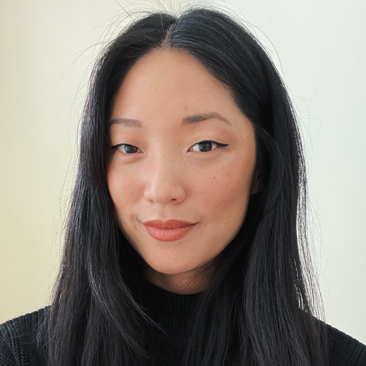 Close-up portrait of a woman with long black hair, fair skin, and brown eyes, wearing a black top, looking at the camera with a slight smile.