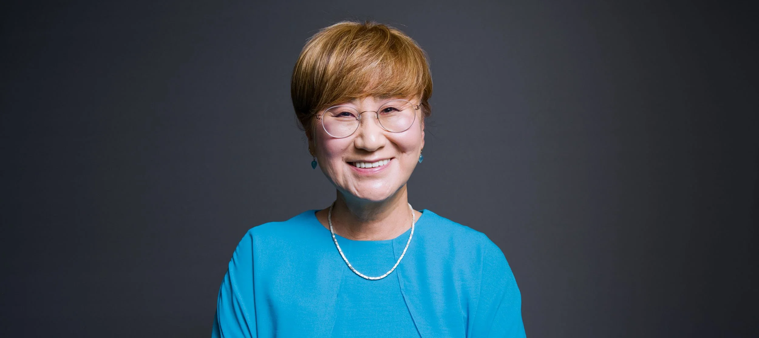 A woman with short red hair, glasses, wearing a blue top and jewelry, smiling at the camera against a dark gray background.