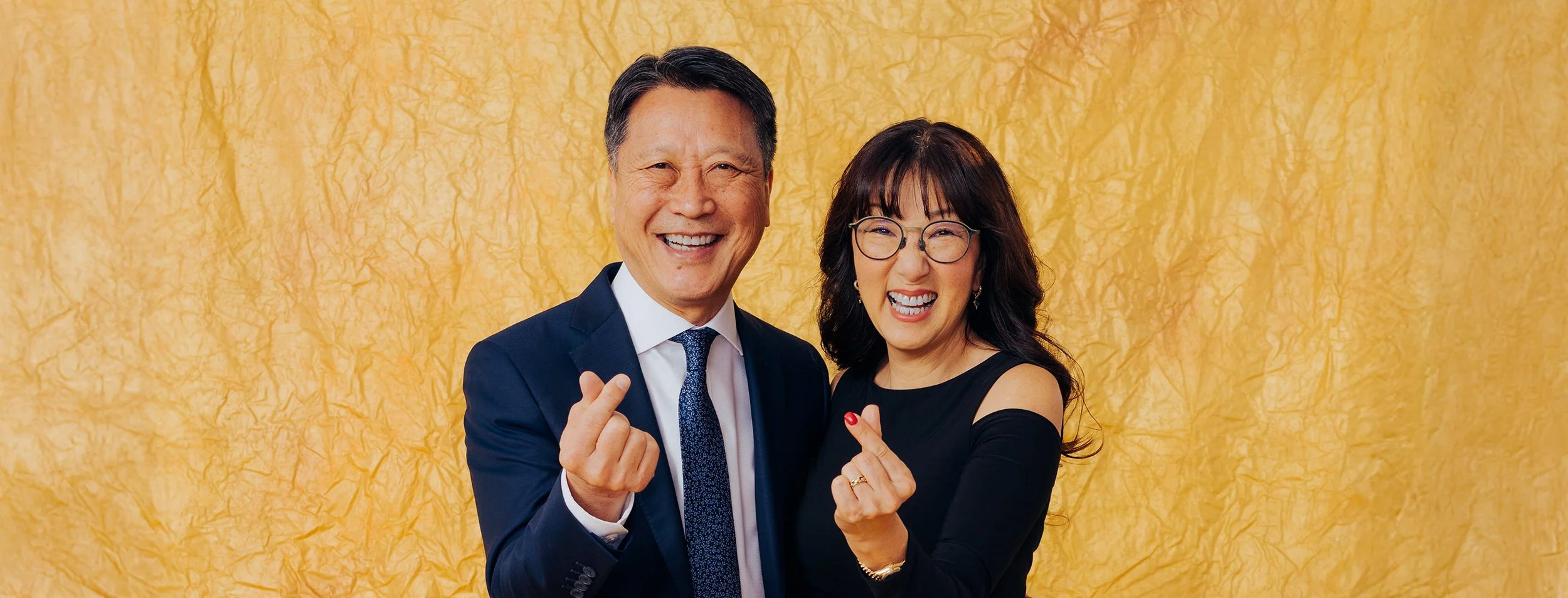A cheerful man in a blue suit and a smiling woman with glasses and dark hair posing against a yellow textured background, both making finger heart gestures.