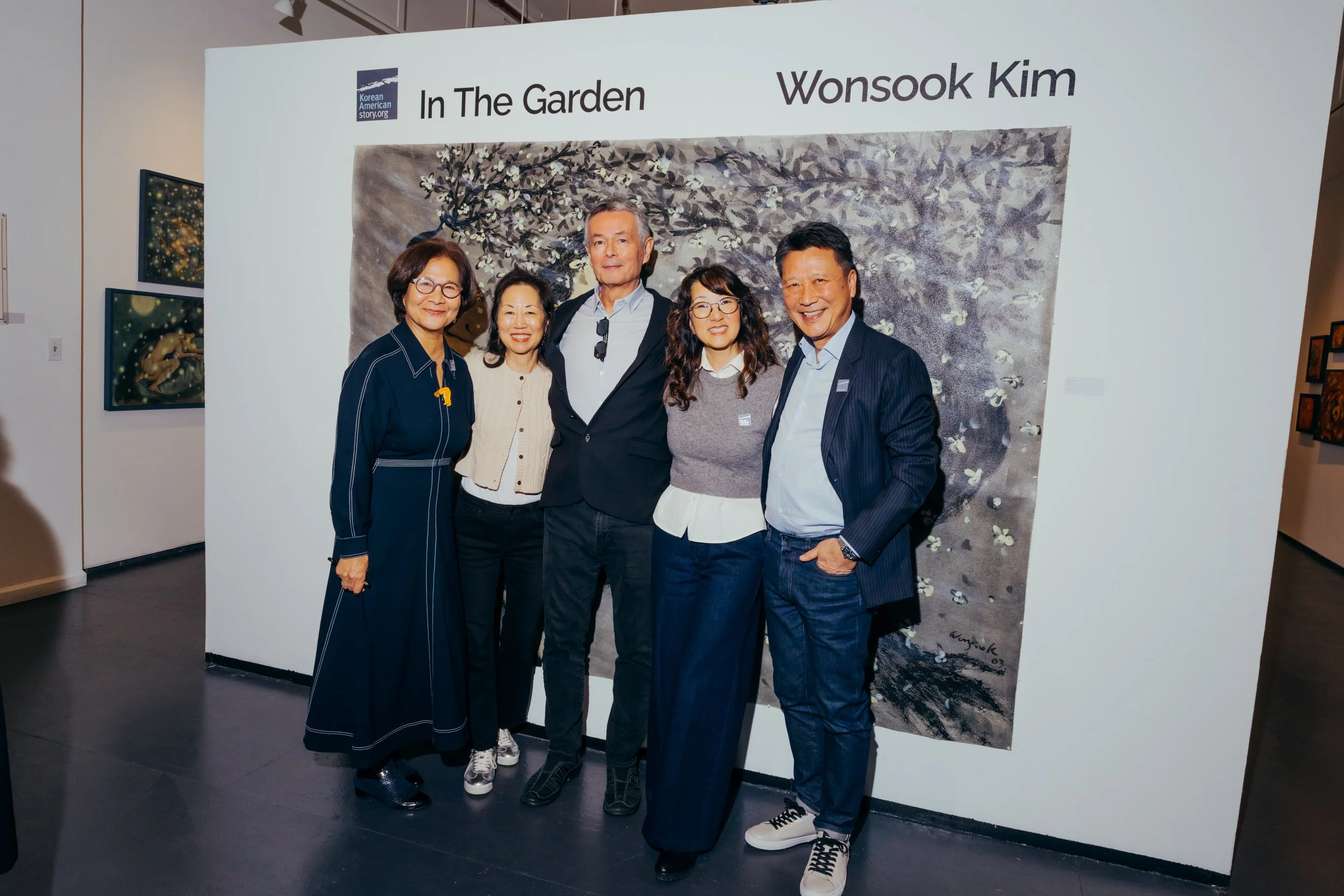 A group of five people, three women and two men, standing together at an art exhibition, posing in front of a large exhibit panel titled 'In The Garden' by Wonsook Kim, with artwork visible behind them.