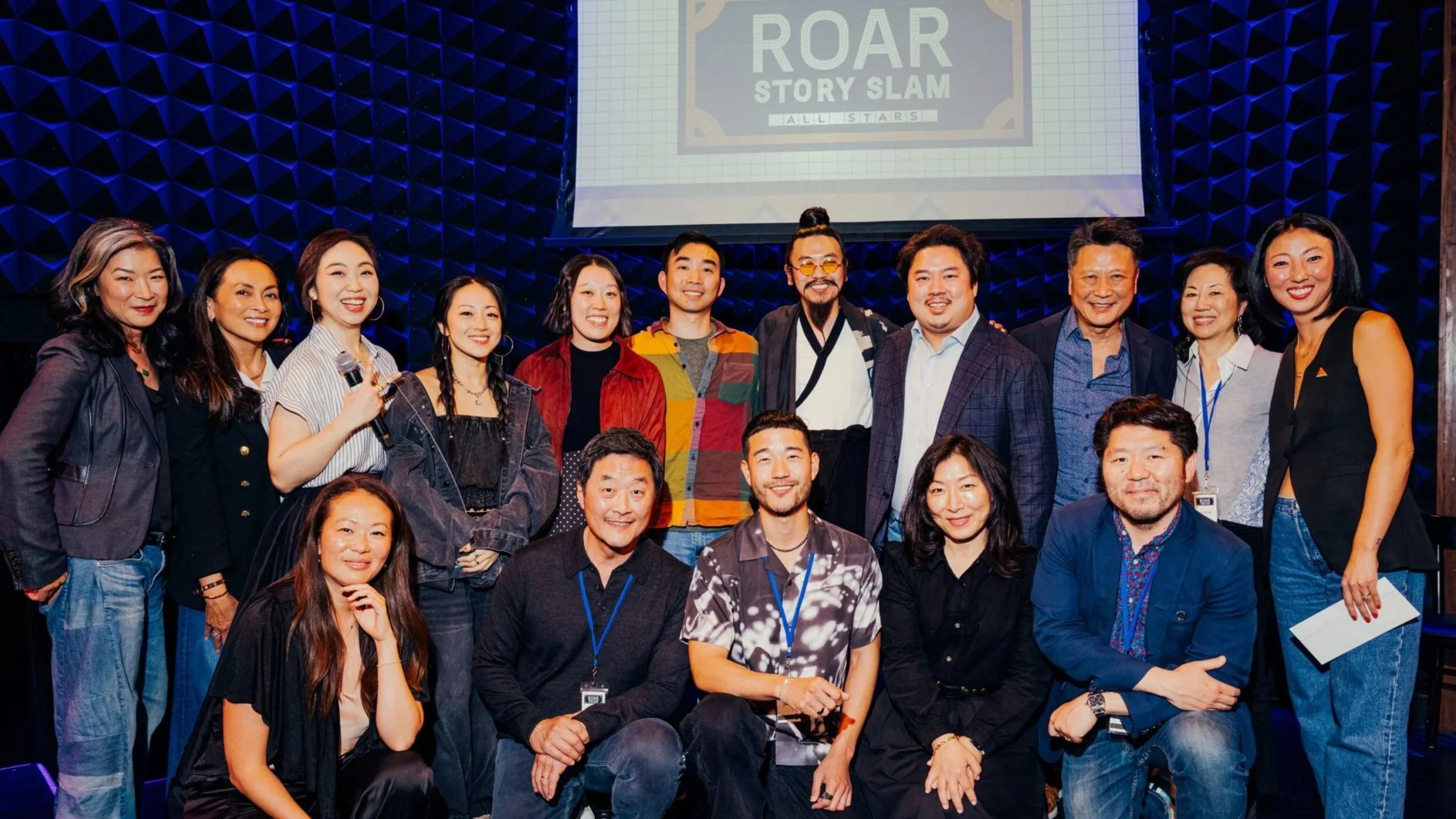 Group of diverse people gathered on stage at an event, with a large screen behind them displaying a sign that says 'ROAR STORY SLAM ALL STARS'.
