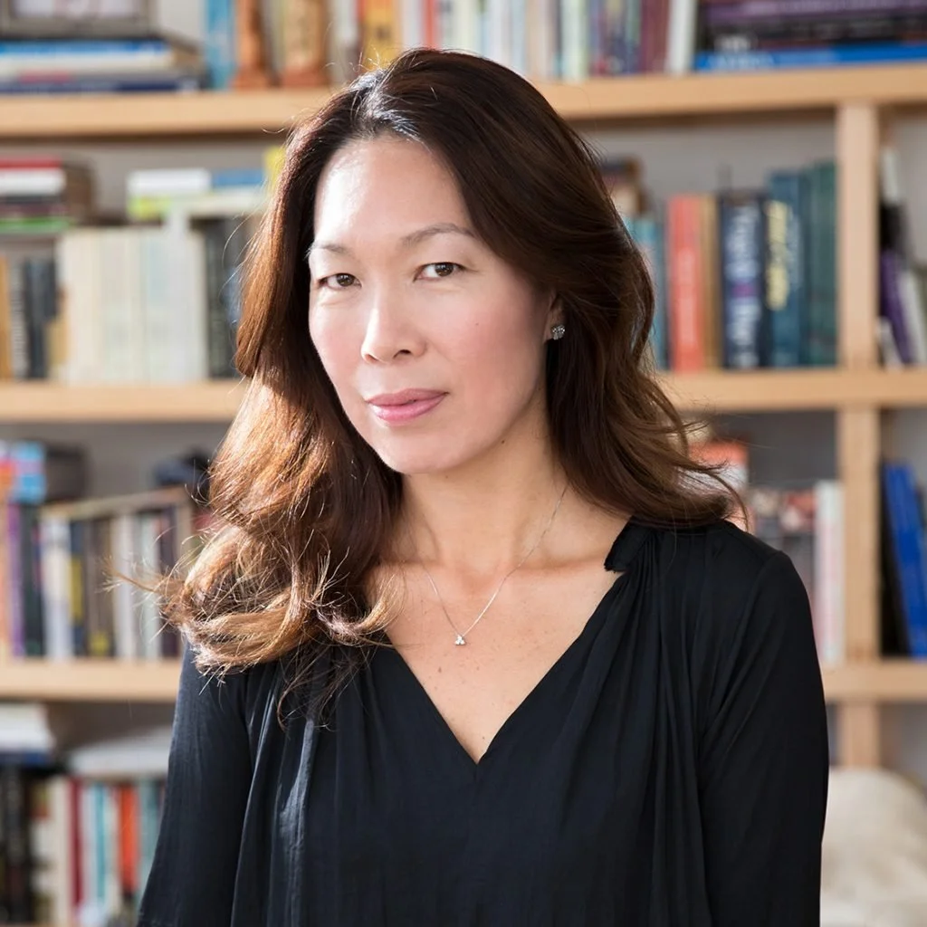 A woman with long, wavy brown hair wearing a black top and a necklace, standing in front of a bookshelf filled with books.