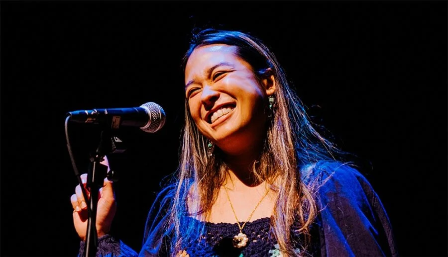 A woman singing or performing on stage with a microphone, smiling with her eyes closed in a joyful expression.