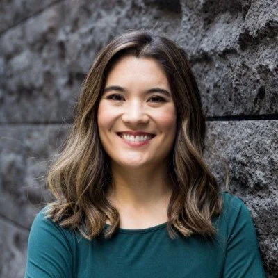 A smiling woman with shoulder-length brown hair standing against a stone wall.