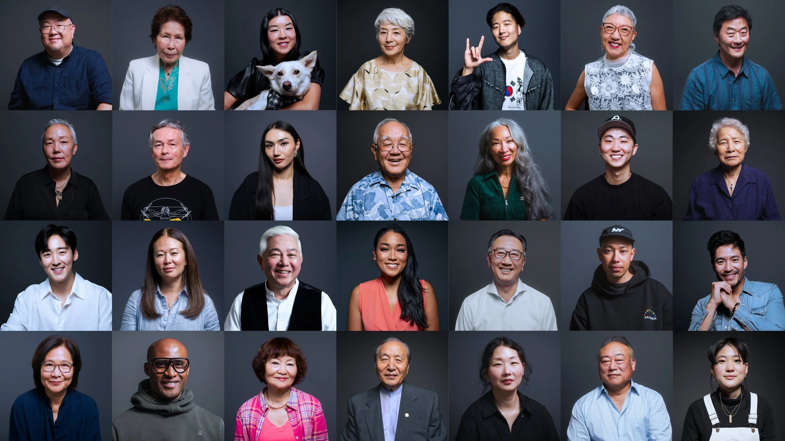 A diverse group of 29 people of various ages, ethnicities, and styles pose against a dark gray background, smiling and looking at the camera.