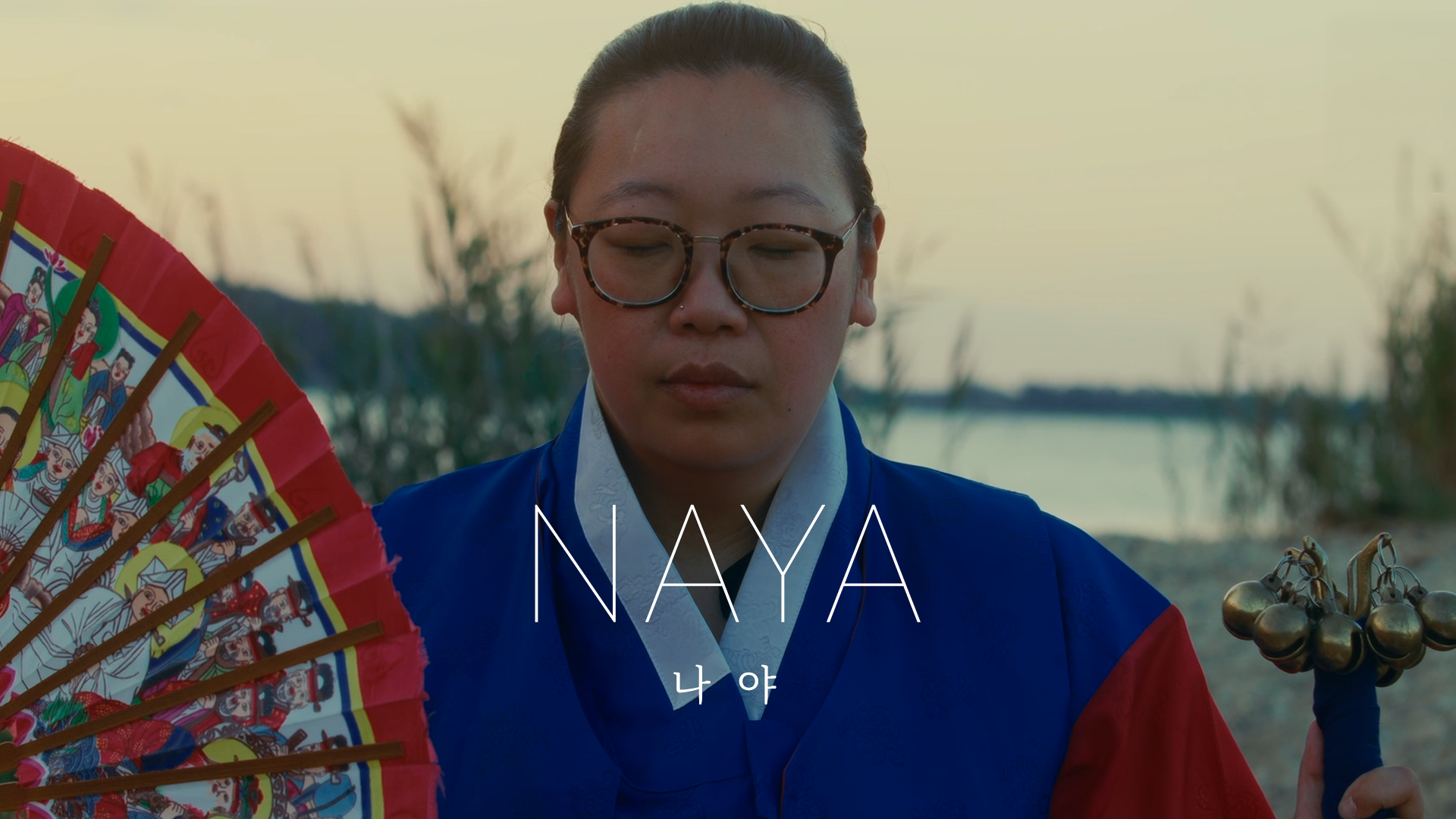 A woman with glasses and a traditional Korean traditional dress, holding a colorful fan and bells, standing outdoors near a body of water during sunset.