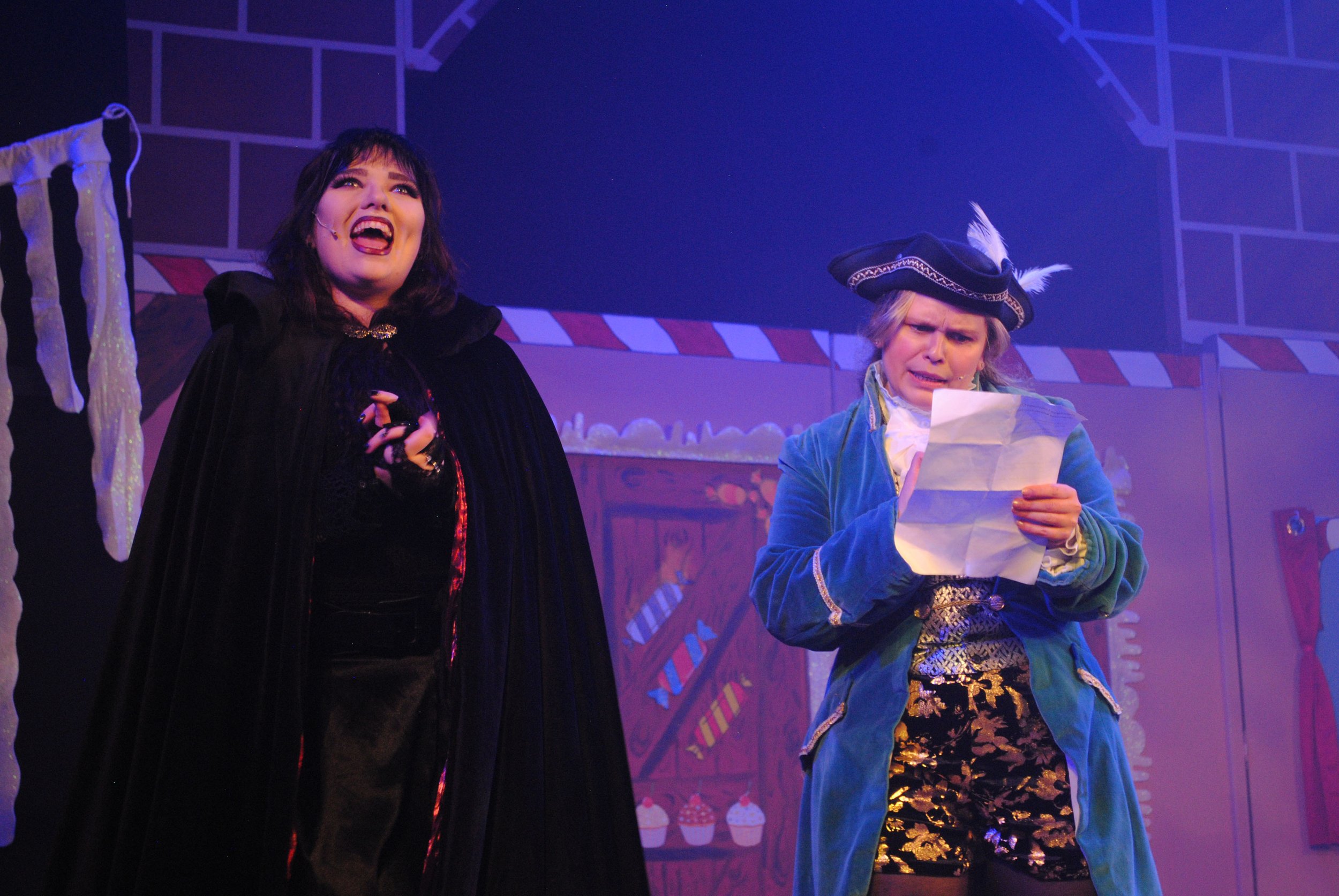 Two women, one dressed as a witch and the other as a pirate, performing on stage with a festive background.