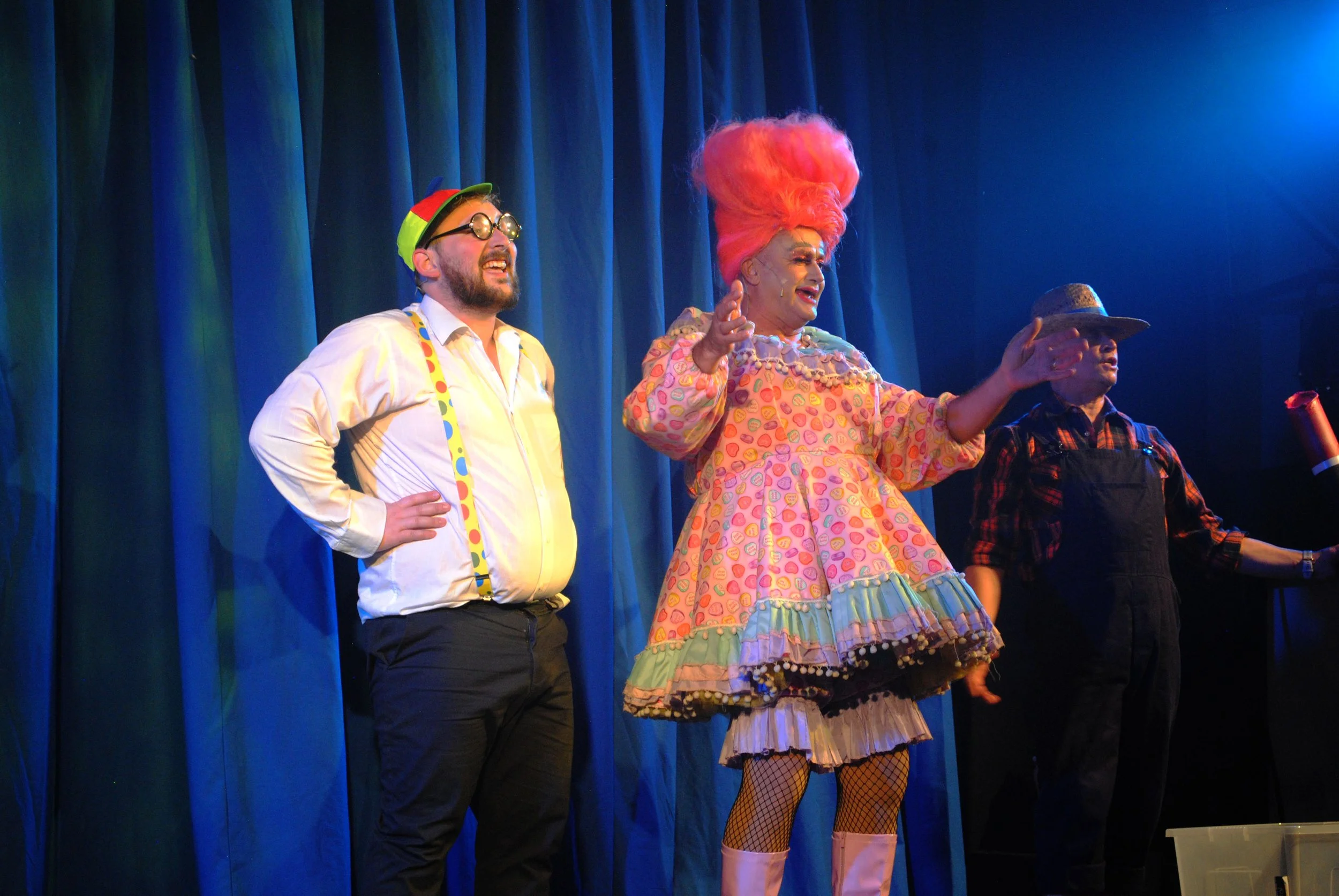 Three performers on stage during a theatrical show with blue curtains in the background. The central performer is dressed in a colorful, eccentric costume with a large pink wig. The performer on the left is wearing glasses, a white shirt with suspend