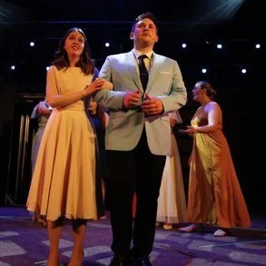 Two people, a woman in a yellow dress and a man in a light-colored suit, standing together at an event with others in the background on a dark stage with string lights.