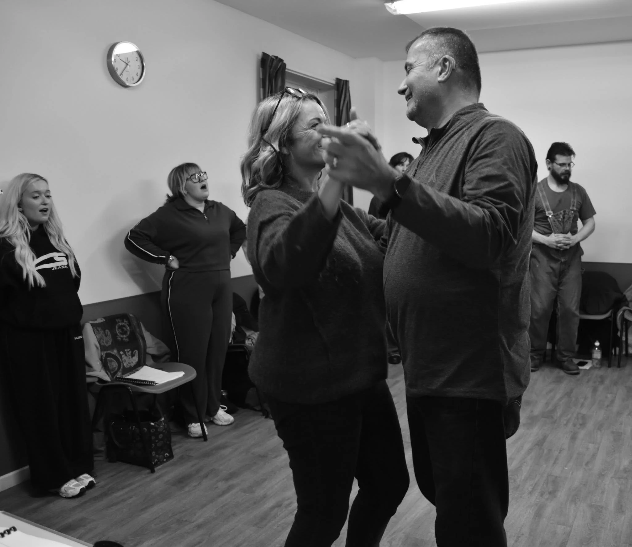 A couple dancing closely together in the center of a room, smiling and looking into each other's eyes, with onlookers in the background.