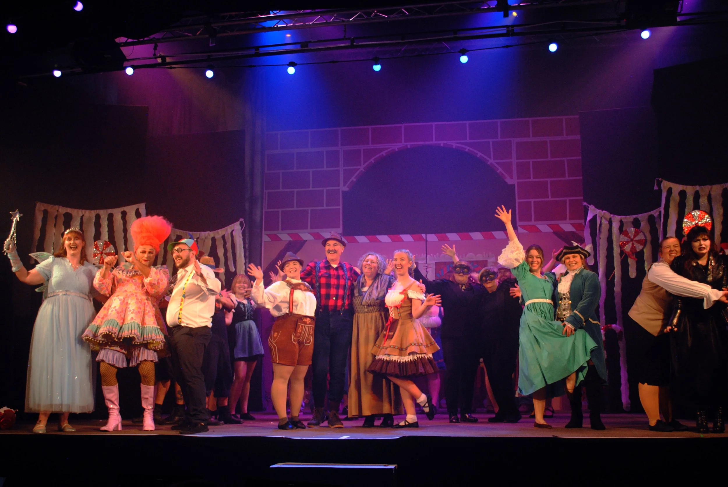 The cast of a theatrical show on stage, dressed in colorful, vintage costumes, smiling and waving to the audience, with a festive gingerbread-themed set in the background.