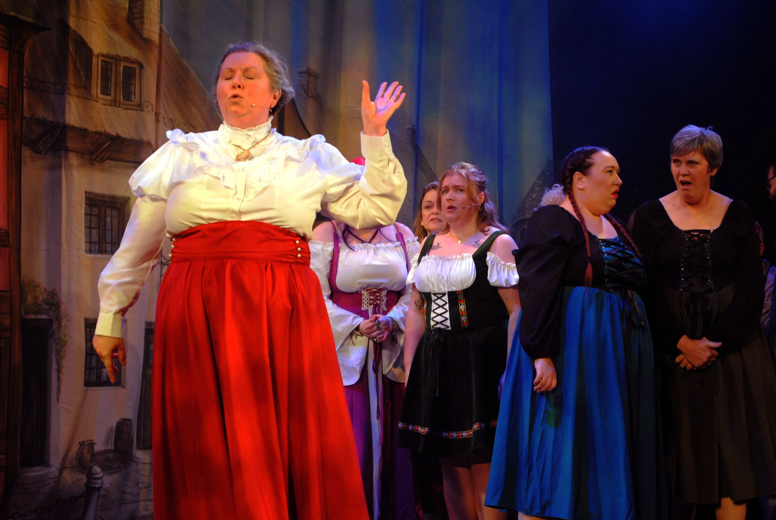 Women dressed in traditional European peasant costumes, some with braided hair, standing on stage during a theatrical performance, with a painted backdrop of buildings.