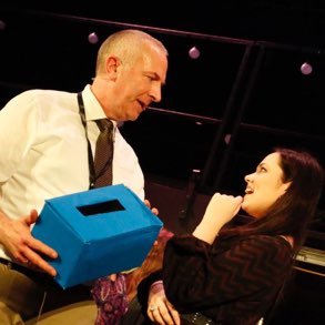 An older man with a bald head and a woman with long dark hair engaging in a conversation, with the man holding a blue box.