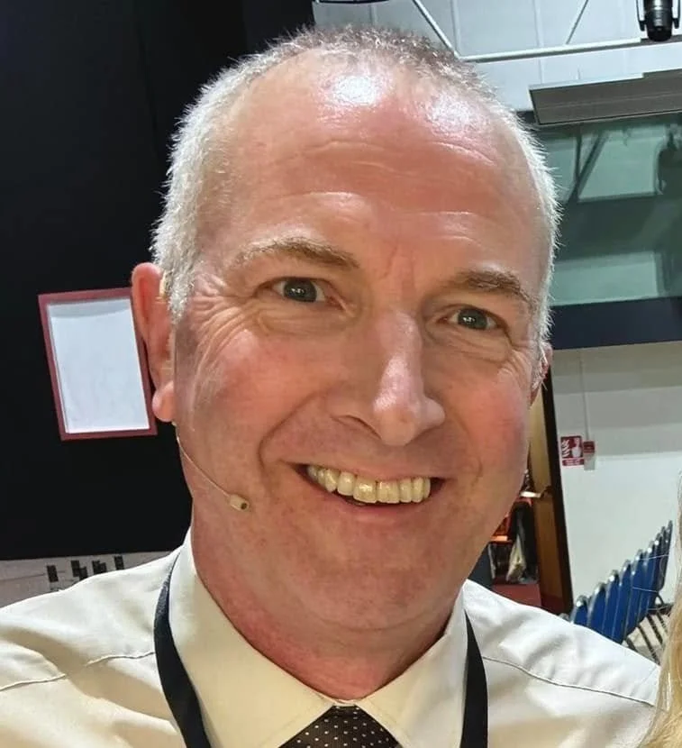 A smiling middle-aged man with short gray hair, wearing a white dress shirt and a dark tie, standing indoors in a conference or event setting.