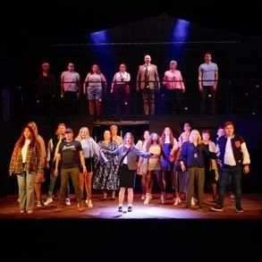 Group on stage with actors and singers, arranged in two rows, with lighting highlighting the performers.