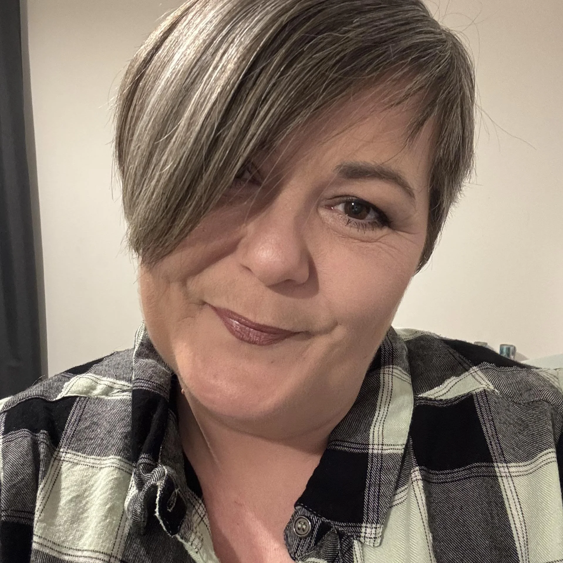 A woman with short hair styled over one eye, wearing a plaid shirt, smiling at the camera.