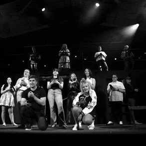 Group of young performers on stage, some sitting and some standing, with a few in the background.