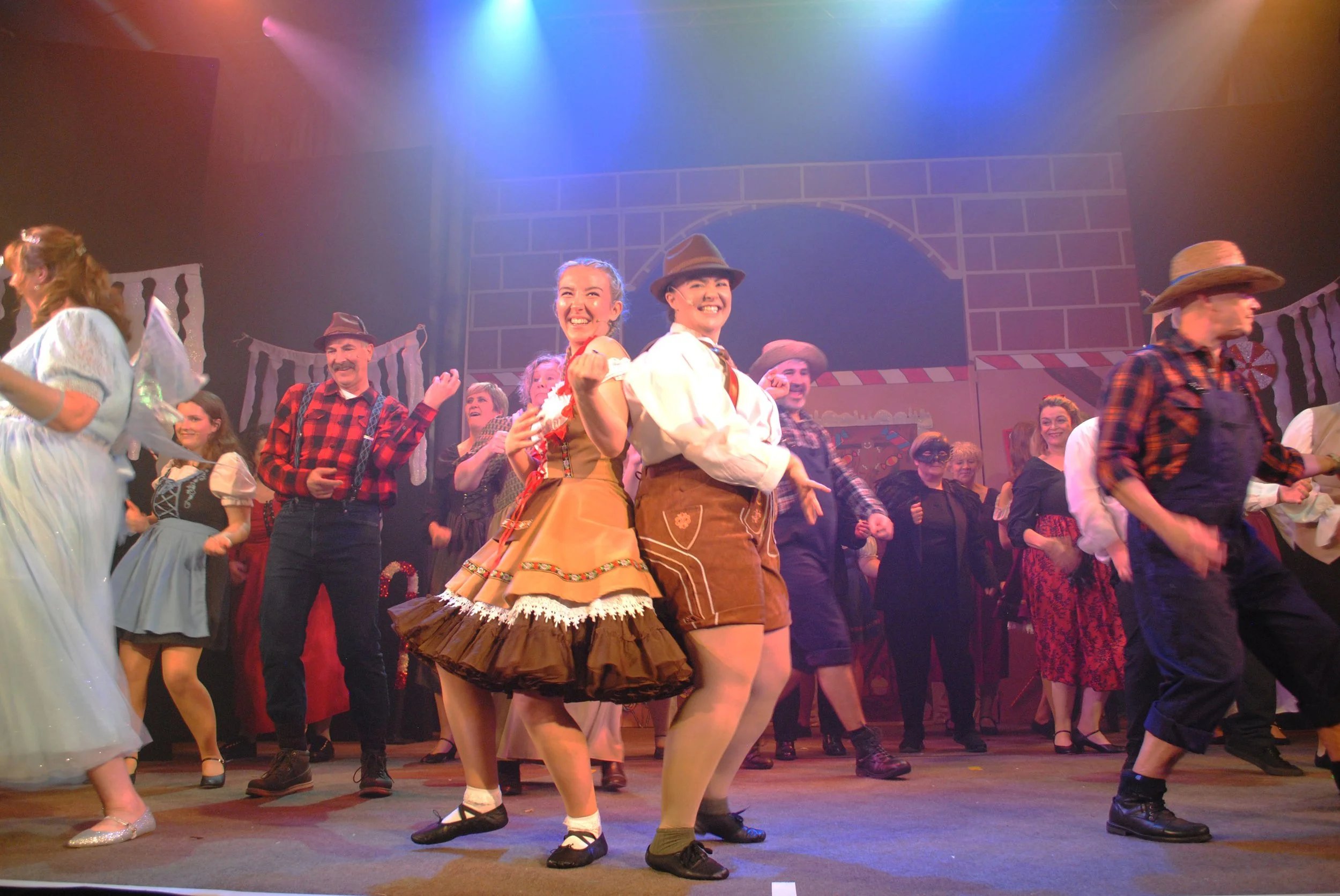 People on stage performing a dance in a theatrical production with colorful lighting and a backdrop resembling a brick arch.