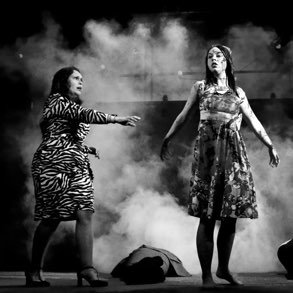 Two women performing on stage with a smoky background, one in a zebra-print dress and the other in a floral dress.