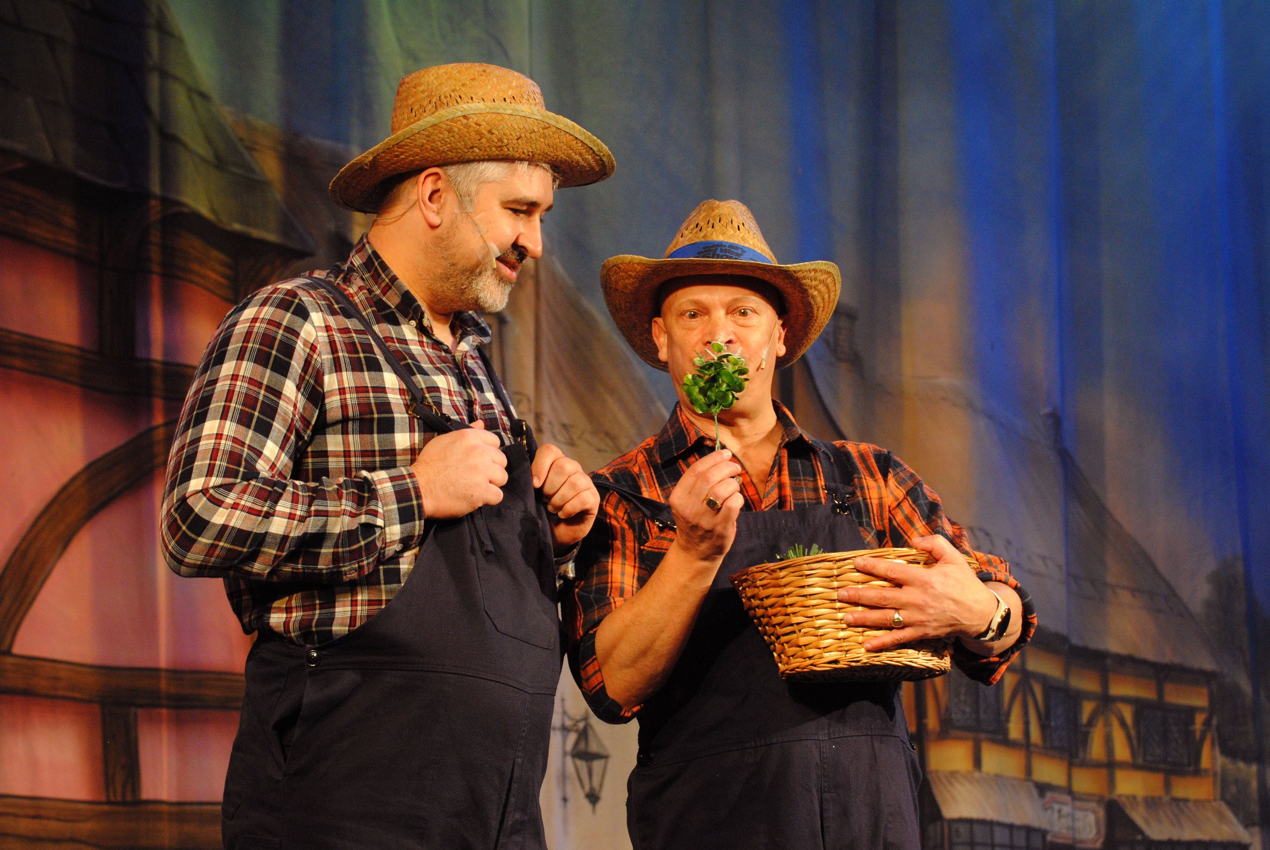 Two men in cowboy hats and plaid shirts, wearing black aprons, sharing a moment on stage with a rustic background. One man is holding a basket and the other a sprig of greenery.