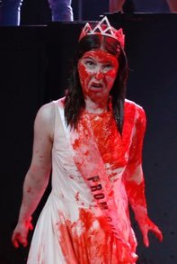 A person dressed as a prom queen, covered in red substance resembling blood, wearing a tiara, with face makeup and a white dress with a sash.