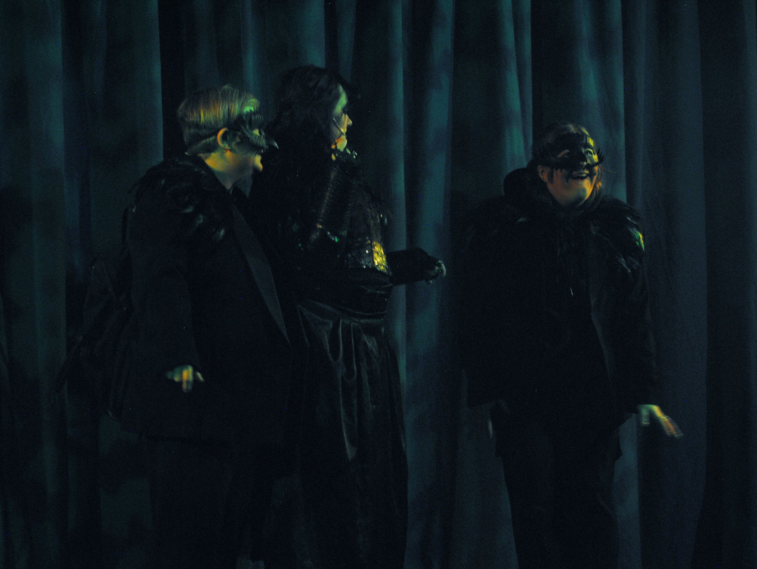 Three women in costumes standing against a dark curtain, appearing to be on a stage, engaged in conversation or performance.