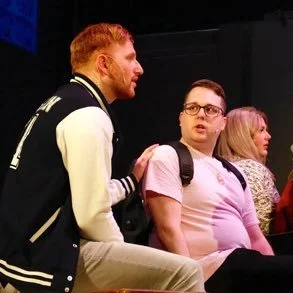 Three people sitting in a row, engaged in conversation. One man with red hair and a beard wearing a black jacket with white sleeves talks to a woman with glasses and a pink shirt. Another woman with blonde hair sits beside them.