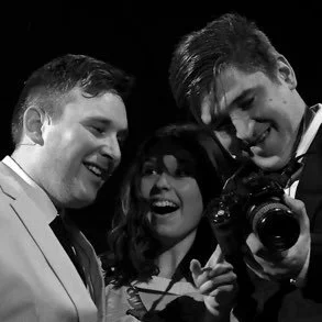 Three people smiling, two men and a woman, with one man holding a camera, in black and white.