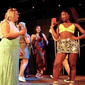 Three women stand on a stage, holding drinks, with two others in the background.