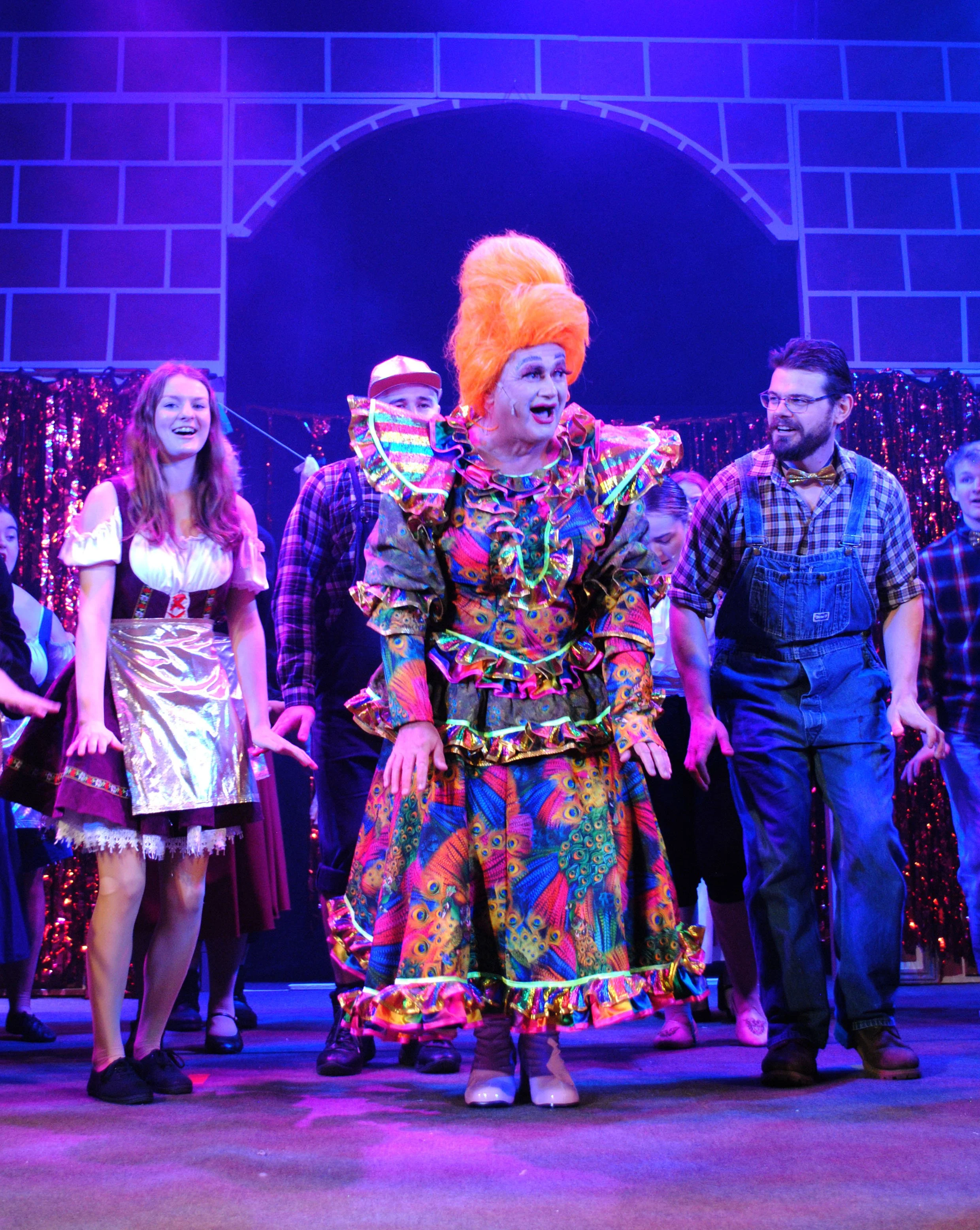 Scene from a theatrical play featuring a woman in a colorful, frilly costume with a large orange wig, surrounded by actors in casual and costume attire on a stage with a brick and purple-lit background.