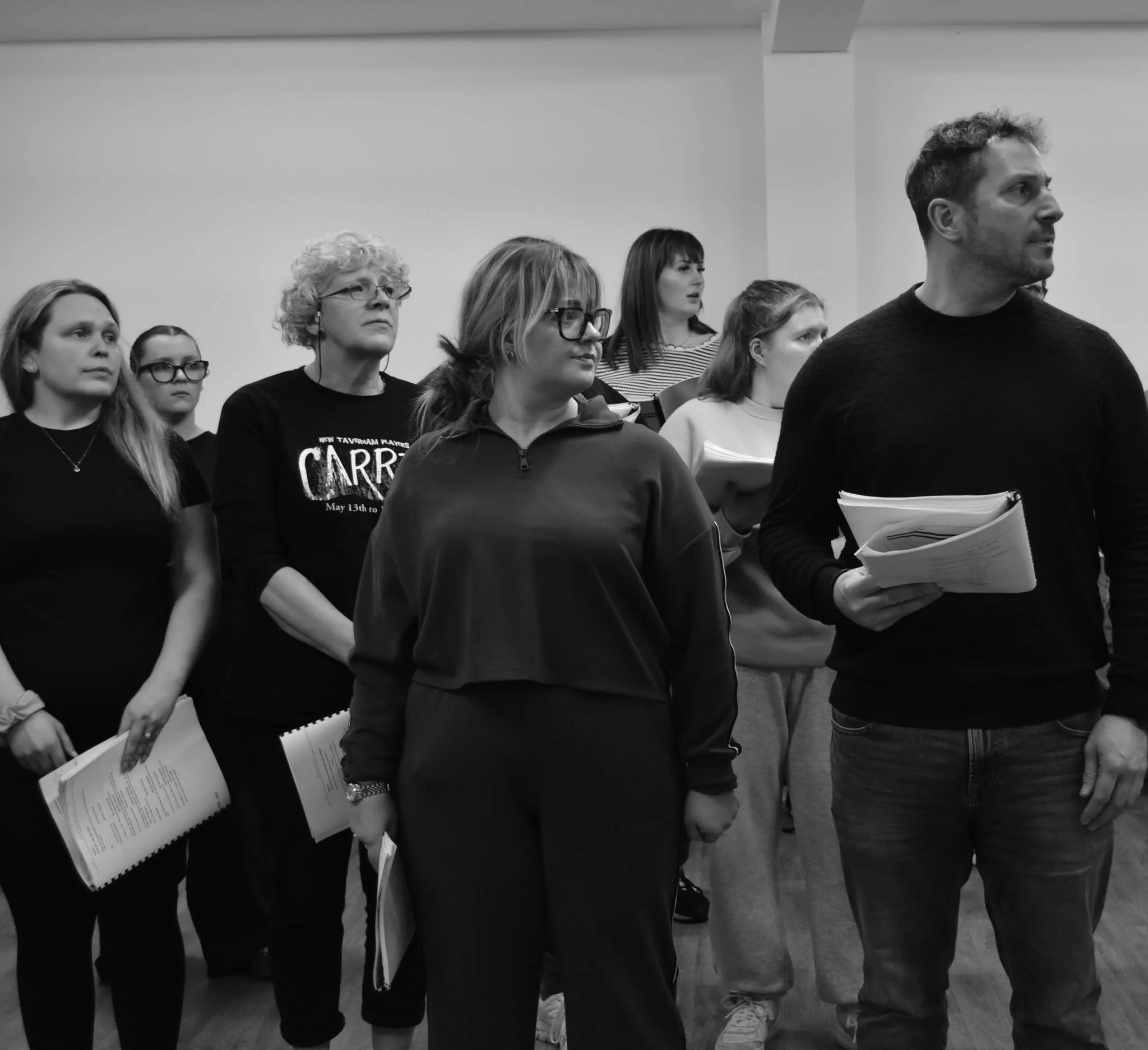 Group of people standing indoors, holding scripts or papers, appearing to rehearse or participate in a rehearsal.