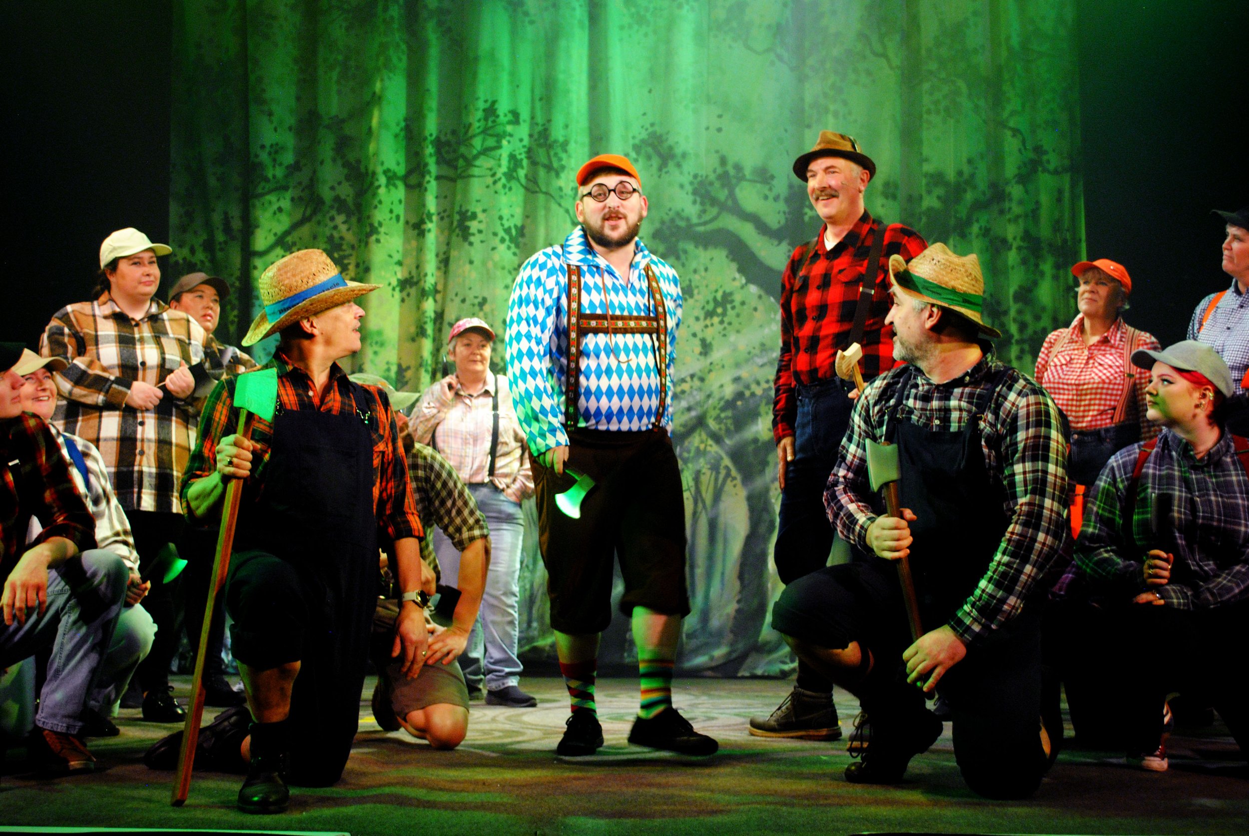 A theatrical scene with a group of actors on stage dressed as forest farmers or lumberjacks, wearing plaid shirts and overalls, set against a backdrop of a green forest. The central figure is a man with glasses, wearing a blue and white diamond-patte