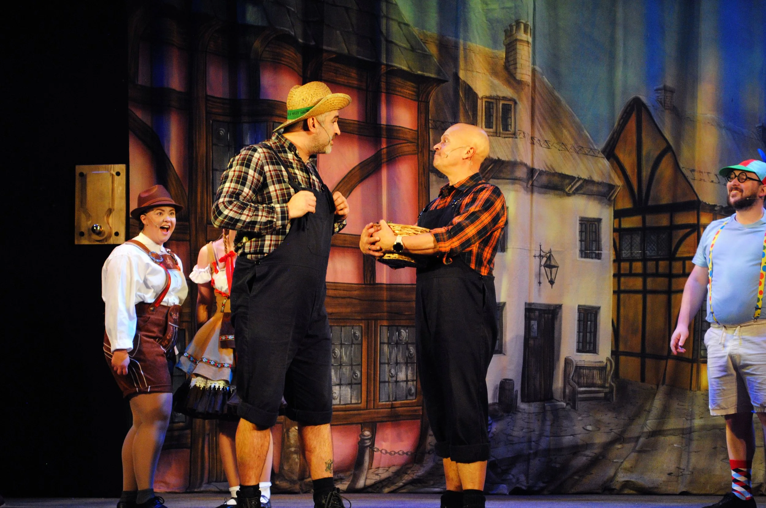 Theater scene with actors in colorful costumes on stage, set against a painted urban background, engaged in dialogue.