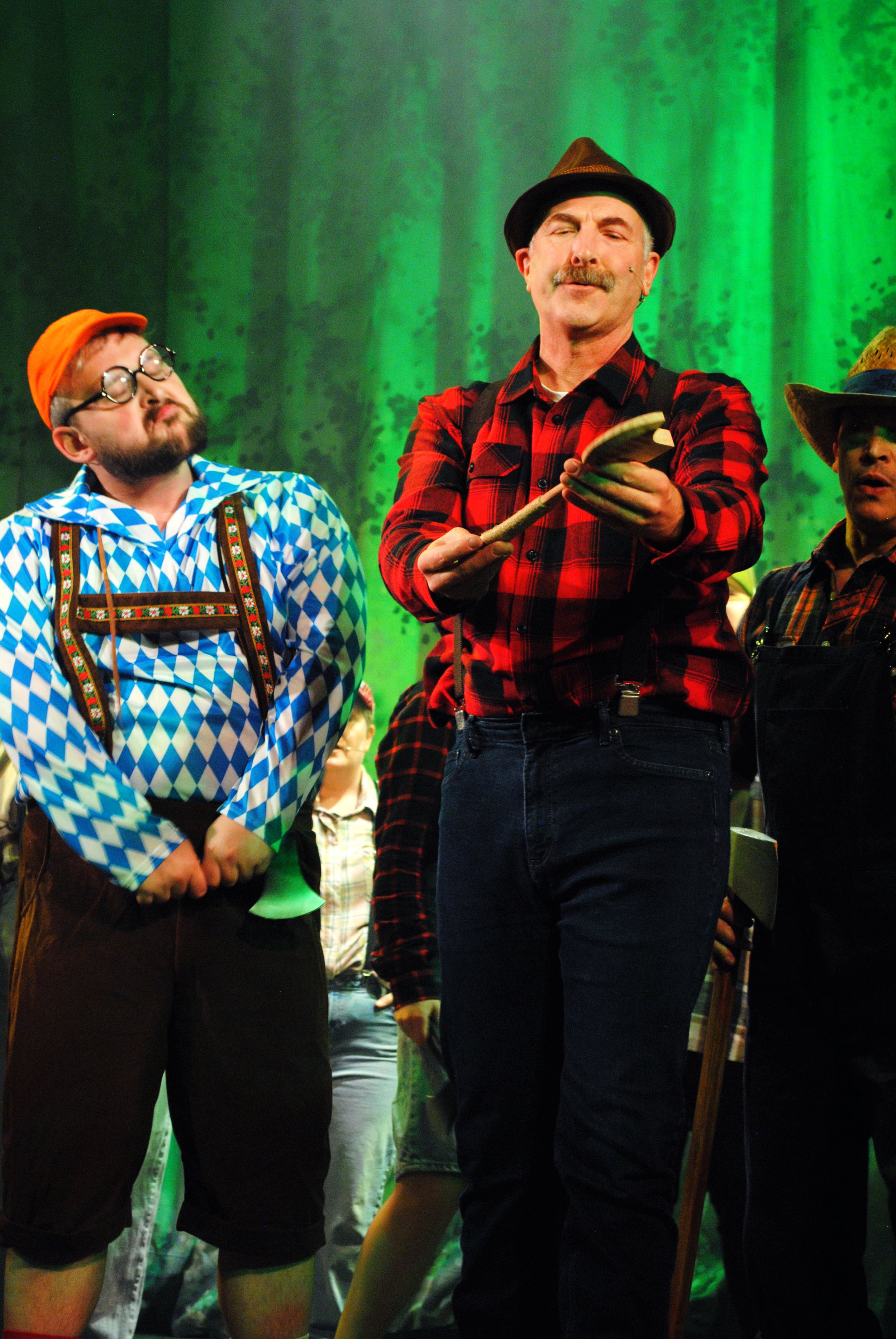 A man in traditional Bavarian attire, including a checkered shirt and suspenders, stands with his eyes closed and hands clasped. Next to him is a man in a red plaid shirt and a hat, holding a fan-shaped object, with a green backdrop behind them.